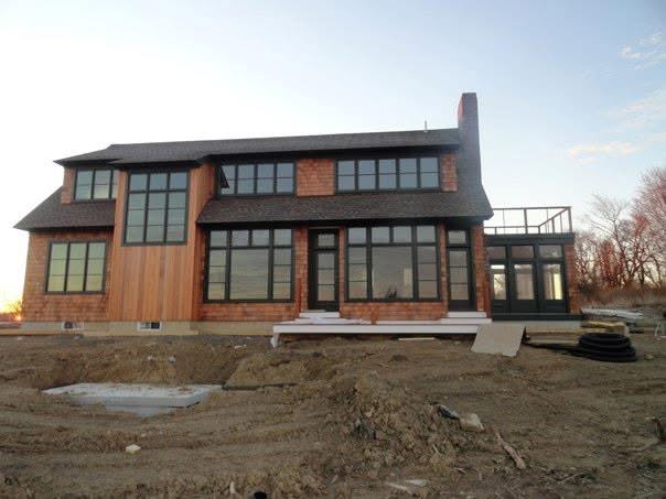 A two-story house under construction, featuring a brown exterior with black-framed windows, a chimney, and a deck. The sur...